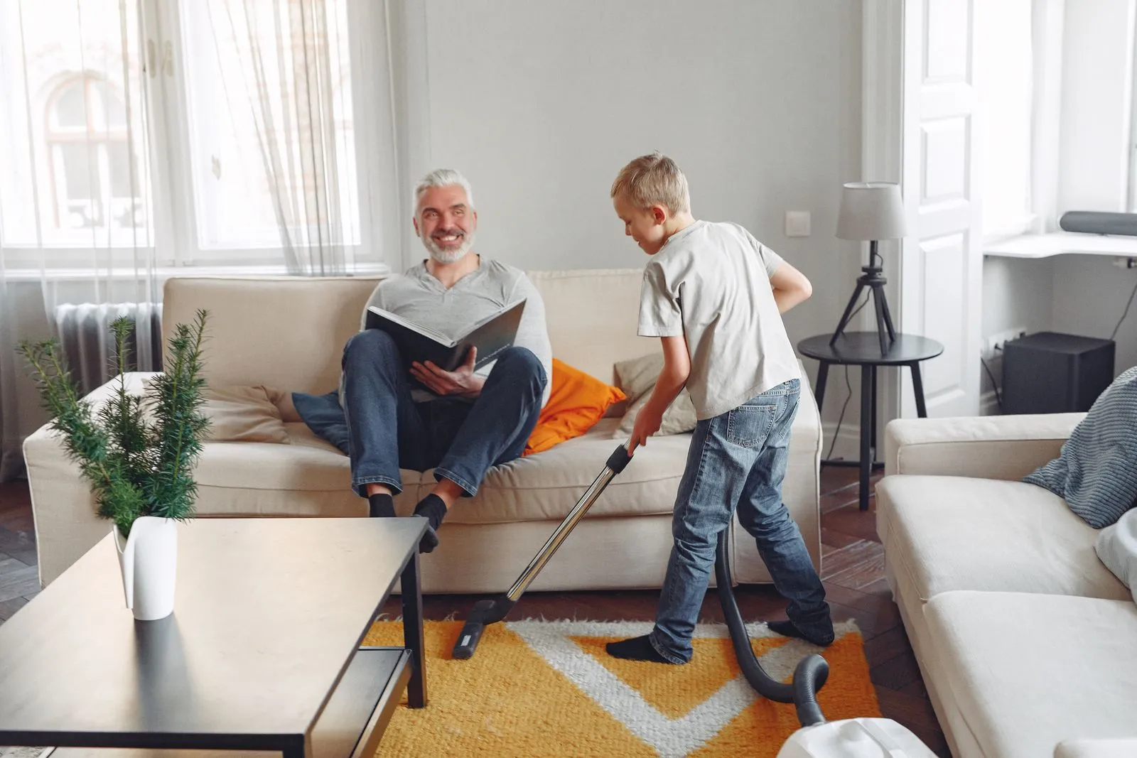 A father relaxing while his son vacuums in a cozy living room setting, showcasing family bonding.